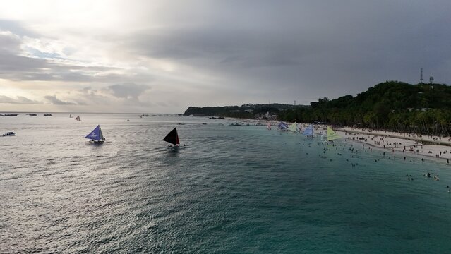 Malay, Philippines - 02 May 2026: Aerial view of Boracay Island with colorful sailboats on turquoise water and people along the white sand beach under a cloudy sky.