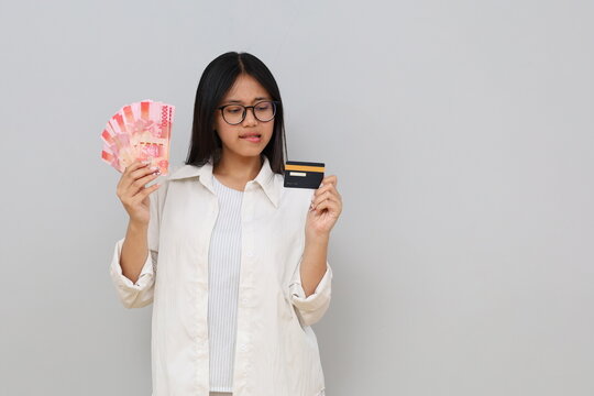 Portrait of an anxious Asian woman holding a credit card and rupiah banknotes, standing in front of a white background.