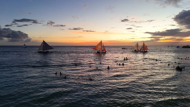 Malay, Philippines - 02 May 2026: Aerial view of Boracay Island at sunset with traditional paraw sailboats on the water and people swimming near the shore under a golden sky.