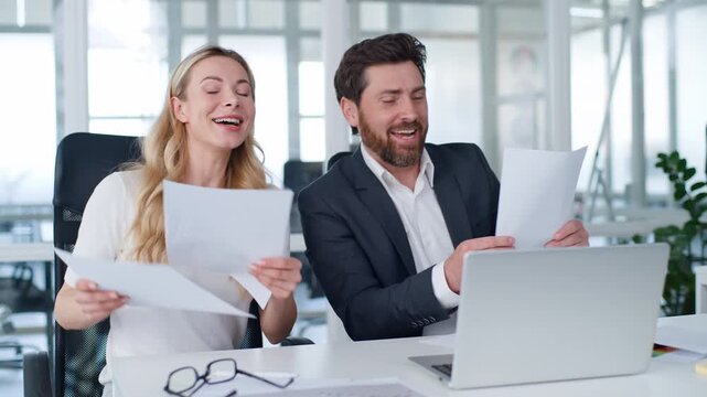 Two Caucasian business executives singing managing papers at office table middle-aged businessman dance sing work with businesswoman enjoy paperwork manage reports happy man with woman dancing working