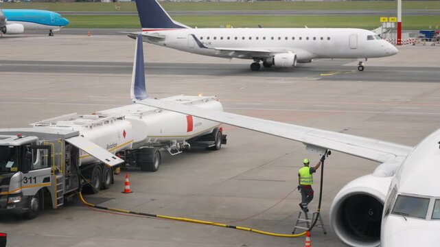 Jet fuel filling process of passenger airplane on airport apron with another commercial airliner taxiing in the background for aviation transport industry traffic logistics service operations.