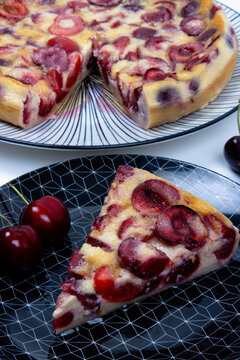 Part de clafoutis aux cerises fait maison sur une assiette noire &agrave; motifs avec des cerises fra&icirc;ches