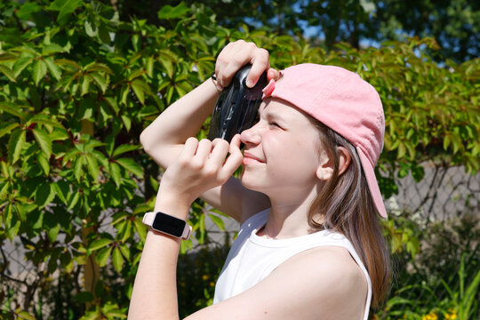 Girl looking through viewfinder of vintage camera outdoors. Enthusiastic tween shooting sunny sky or green trees on retro film cam. Creative young photographer exploring nature photography during