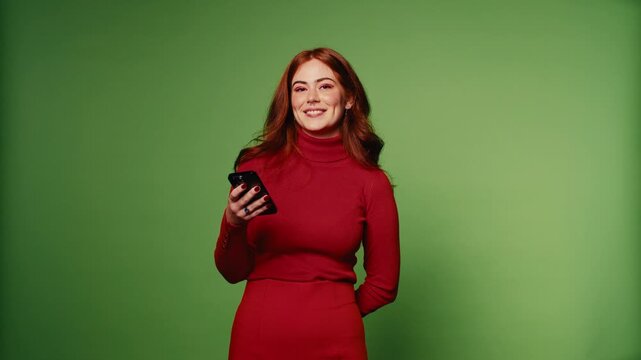 A happy redhead woman in a red dress uses her phone against a vibrant green screen background.