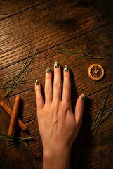 Fototapeta premium Female hand displaying trendy green and gold checkered nail art on a rustic wooden table with pine needles, cinnamon, and dried orange.