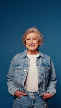 Happy senior woman in denim jacket and jeans smiles and laughs on blue studio background. Vertical video.