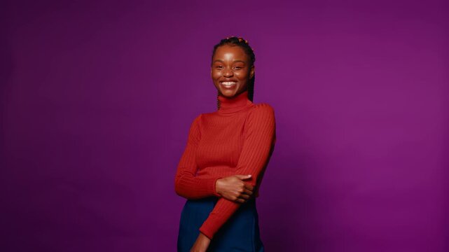 Relaxed young woman stands against a vibrant purple backdrop, her smile radiating positivity and joy as she looks towards the camera.