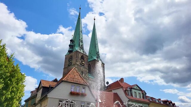 Blick &uuml;ber einen springenden Brunnen auf historische Fachwerkh&auml;user und die markanten Doppelt&uuml;rme der St. Nikolai Kirche in Quedlinburg unter einem blauen Sommerhimmel mit Wolken.
