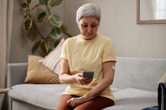 Senior woman checking smartphone while monitoring glucose sensor on arm at home, managing diabetes and digital health routine. Useful for telehealth, chronic care, wellness campaigns