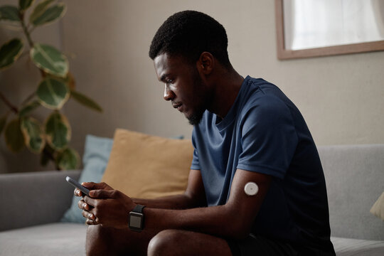 Young Black man checking smartphone while monitoring glucose sensor on arm, managing diabetes at home and tracking health data. Side view, ideal for telehealth, wellness, chronic care marketing