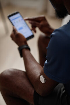 Vertical shot of Black man checking glucose data on smartphone while wearing continuous monitor, managing diabetes care. Ideal for telehealth, health app, patient education