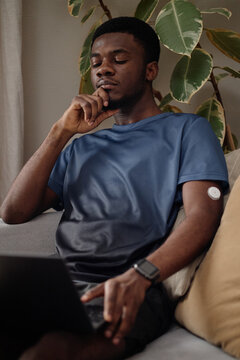 Vertical shot of young Black man wearing glucose sensor, using laptop and touching chin while monitoring health data at home. Supporting diabetes management, telehealth, wellness marketing
