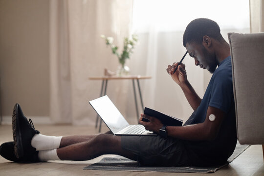 Young African American man with glucose sensor sitting on floor, using laptop and taking notes, managing diabetes and remote routine. For telehealth, chronic care, wellness campaigns