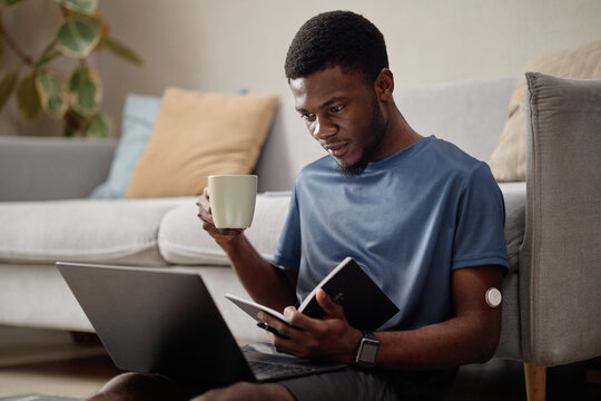Young Black man with insulin pump on arm working on laptop while holding notebook and coffee, managing home tasks. Ideal for remote work, productivity, freelance lifestyle marketing
