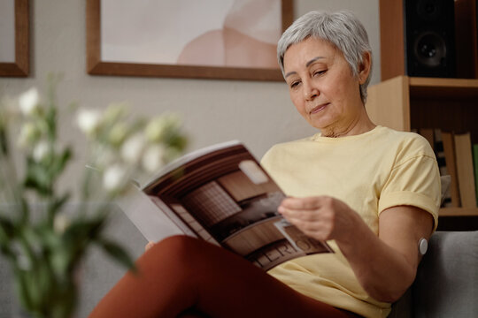Senior Asian woman reading magazine while sitting at home, showing glucose sensor on arm and independent diabetes management. Useful for healthcare campaigns, patient education