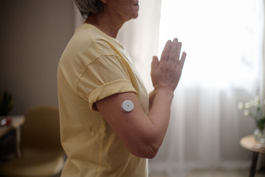 Senior Caucasian woman wearing glucose sensor and practicing yoga at home, representing diabetes management and healthy aging. Useful for healthcare, wellness, remote monitoring campaigns