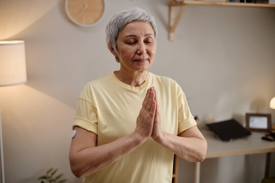 Asian senior woman practicing meditation with hands in prayer pose, closing eyes at home and supporting mindfulness routine. Useful for wellness, stress relief, healthy aging marketing