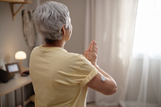 Senior Asian woman practicing mindfulness exercise at home while wearing glucose monitor on arm, representing diabetes self care. Useful for wellness, aging, healthcare campaigns