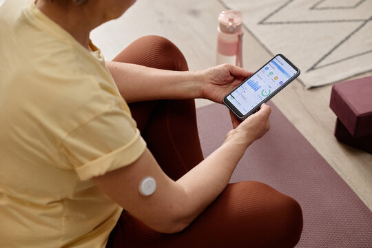 High angle of  woman checking glucose app on smartphone while resting on exercise mat, wearing sensor for diabetes tracking. Ideal for healthcare tech, wellness, self management
