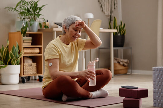 Senior Asian woman resting on yoga mat, wiping forehead and holding water bottle after home workout, showing fitness fatigue and glucose monitoring. Ideal for wellness, diabetes, active aging