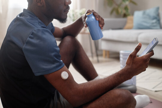 African American man checking smartphone fitness app while resting with water bottle, wearing glucose sensor after workout. Useful for diabetes tech, wellness, remote care marketing
