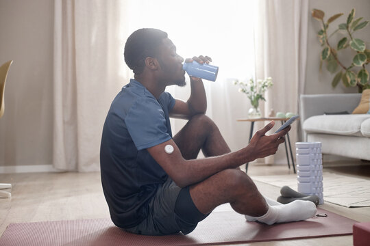 Young Black man resting on exercise mat, drinking shake and checking smartphone after home workout, wearing glucose sensor. Ideal for fitness app, diabetes management, wellness marketing