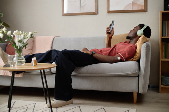 Young adult Black man resting on sofa while checking smartphone and listening through headphones, showing diabetes management at home. Useful for healthcare, chronic condition, wellness marketing