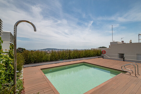 A modern rooftop swimming pool surrounded by greenery. The pool has a wooden deck and a ladder. The sky is clear with a few clouds.