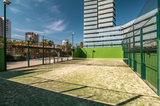 A modern padel court surrounded by urban buildings. The court features a green playing surface and is enclosed by a fence. Clear blue sky above.