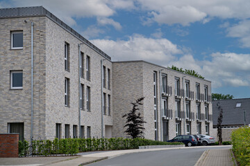 Naklejka premium Exterior view of a contemporary multi-story residential building featuring light brick walls, large windows, and small balconies under a cloudy sky.