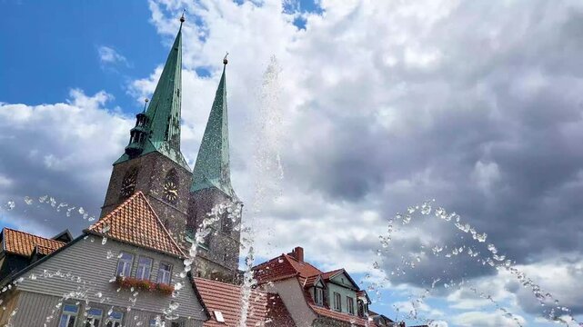 Blick &uuml;ber einen springenden Brunnen auf historische Fachwerkh&auml;user und die markanten Doppelt&uuml;rme der St. Nikolai Kirche in Quedlinburg unter einem blauen Sommerhimmel mit Wolken.