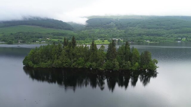 Eilean Gorm Loch drone descent reveals a tree-covered island mirrored on dark water, with misty Highland hills and rural shoreline in Scotland under overcast skies.