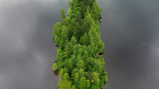 Eilean Gorm, Loch Ard drone shot reversing and tilting up to reveal the full wooded island, dark loch waters and forested shoreline in Loch Lomond and The Trossachs, Scotland.