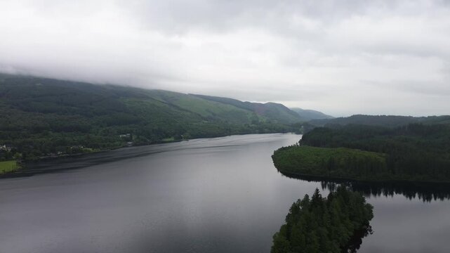 Reverse drone flight over Loch Ard in the Trossachs, Scotland, pulling back above dark water, pine forest shoreline and distant hills under changing light for travel and nature scenes.