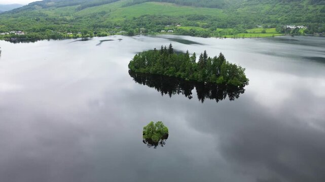 Eilean Gorm on Loch Ard rises into view as a drone ascends above a tree-covered island, reflective freshwater loch, and green Scottish Highlands under overcast skies.