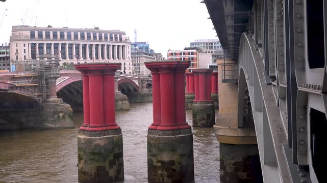 Weathered red bridge pillars stand in the Thames beside the metal structure of Blackfriars rail bridge on an overcast day.