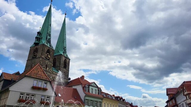 Blick &uuml;ber einen springenden Brunnen auf historische Fachwerkh&auml;user und die markanten Doppelt&uuml;rme der St. Nikolai Kirche in Quedlinburg unter einem blauen Sommerhimmel mit Wolken.