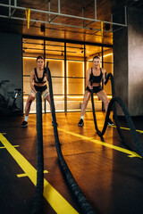 Fototapeta premium Two athletic women performing battle rope exercises in a modern gym with wooden flooring and illuminated background, showcasing strength and fitness training