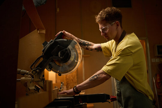 Young adult craftsperson operating miter saw in carpentry workshop, cutting wood for custom production and skilled trade concept. Useful for vocational training, maker business marketing