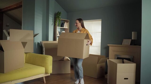 Smiling young woman happily carrying a large cardboard box, unpacking belongings in the living room of her new apartment, starting a new chapter in life and celebrating relocation day