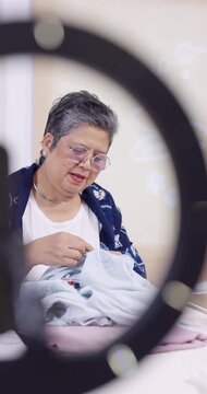Senior Asian woman artisan bends toward denim bundle, stitches resist pattern with red thread, answers live chat, ringlight camera nearby, slow fashion content for home studio small business.