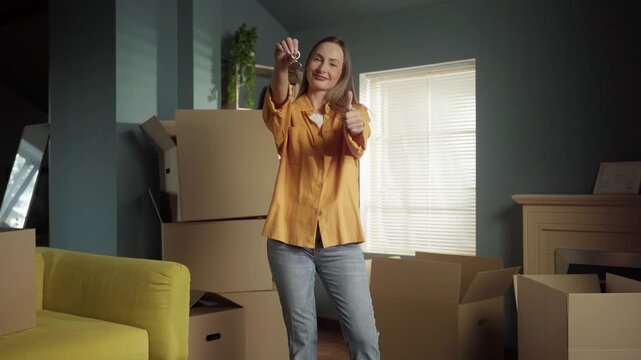 Proud woman standing in her new home among cardboard boxes, holding up the keys to her apartment. Cheerful female renter showing a thumbs up, celebrating a successful real estate deal