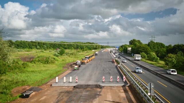Footage of road construction machinery lined up on newly paved carriageway with adjacent traffic flowing on open motorway. Highway expansion and road construction with heavy machinery and traffic. 