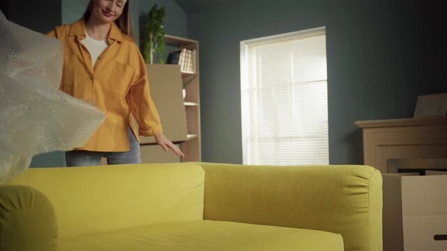 Happy young woman unwrapping a modern yellow sofa in her new apartment's living room, smiling as she removes the protective bubble wrap after moving in and surrounded by cardboard boxes