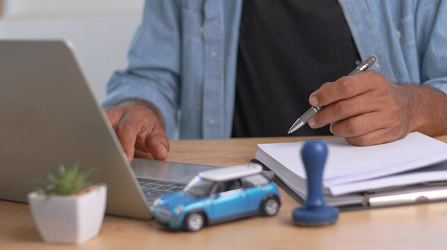 Close-up of a professional reviewing vehicle insurance or auto loan documents while working on a laptop. Car model, paperwork, and office desk symbolize automotive finance, registration, and business 