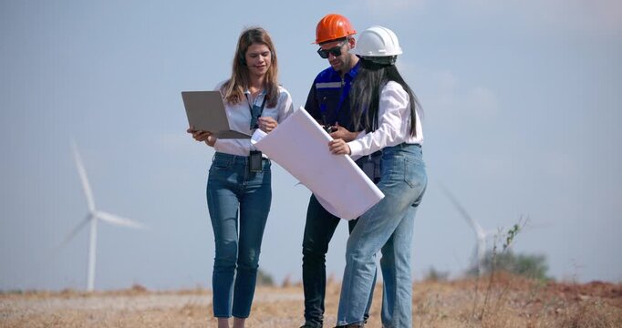 A team of three engineers in hardhats and safety vests review blueprints and discuss a renewable energy project while inspecting wind turbines and planning wind farm development outdoors.
