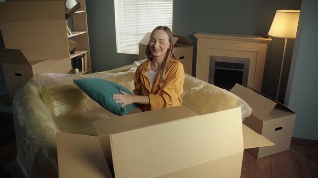 Joyful young woman sitting on a couch covered in bubble wrap, happily unpacking personal belongings, a lamp, and a pillow from a large cardboard box after moving into a new apartment