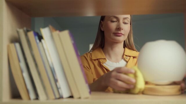 Young woman meticulously wiping dust from a wooden bookshelf and books with a yellow cloth, tidying up the living room during her daily cleaning routine and maintaining a neat home environment