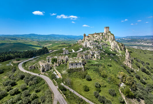 Aerial drone view of the abandoned ghost village of Craco in Basilicata, Italy, showing dramatic hilltop ruins, winding road, green valleys and rugged cliffs under a clear blue sky