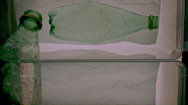 Silhouetted hand placing a crumpled green plastic bottle in a fridge for recycling or reusing